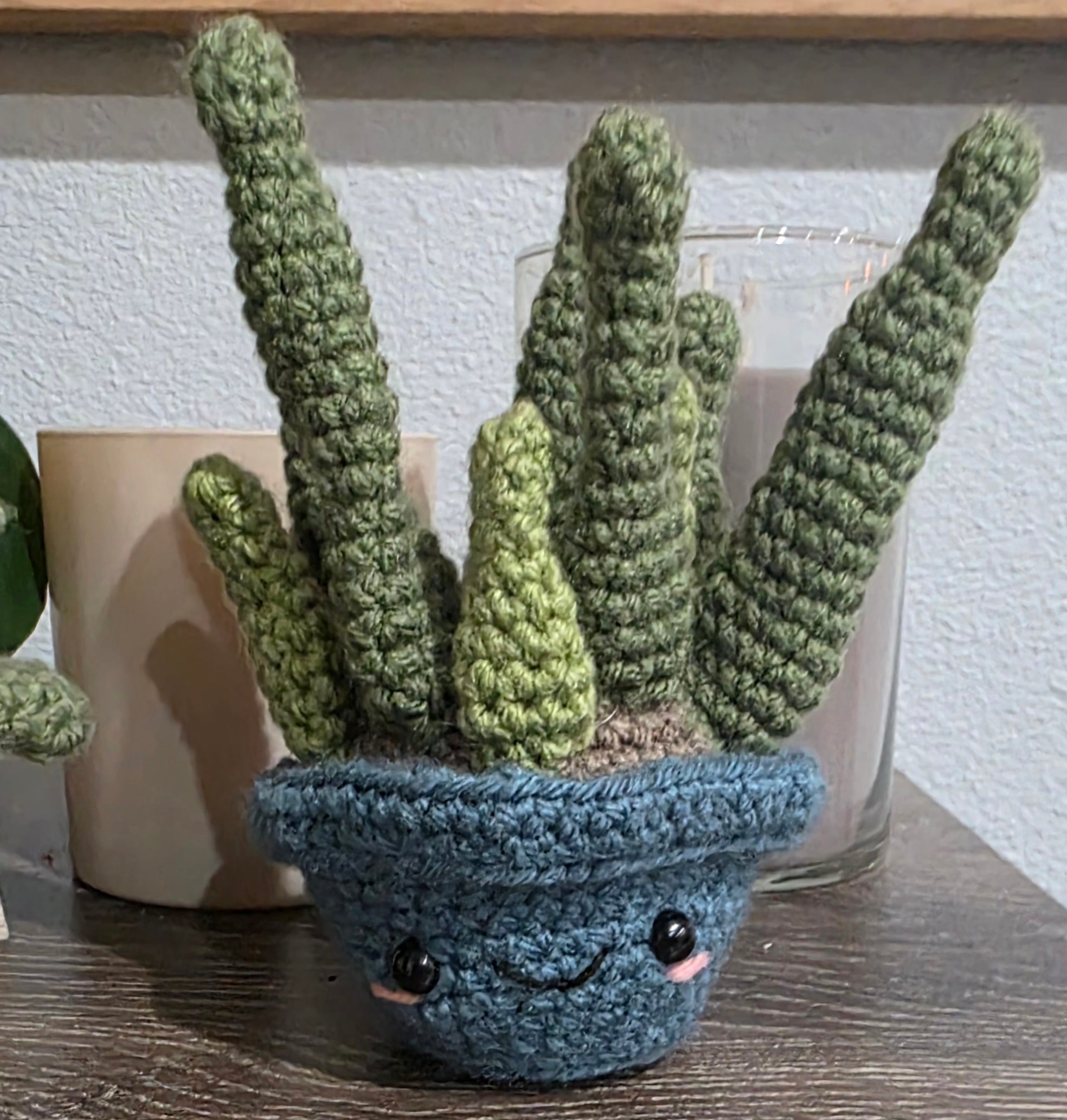 Crocheted aloe plant in a blue pot with a face on a wooden surface.