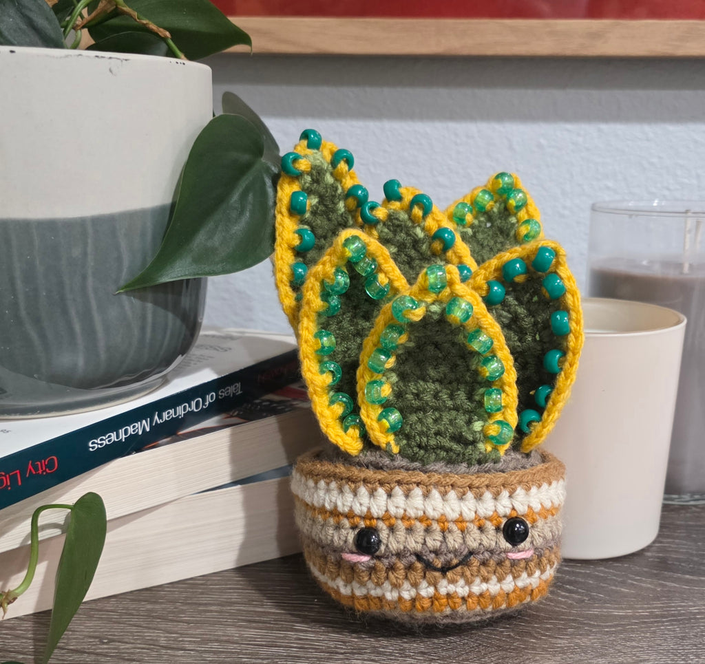 Crocheted potted plant with a face on a table with books and a glass.