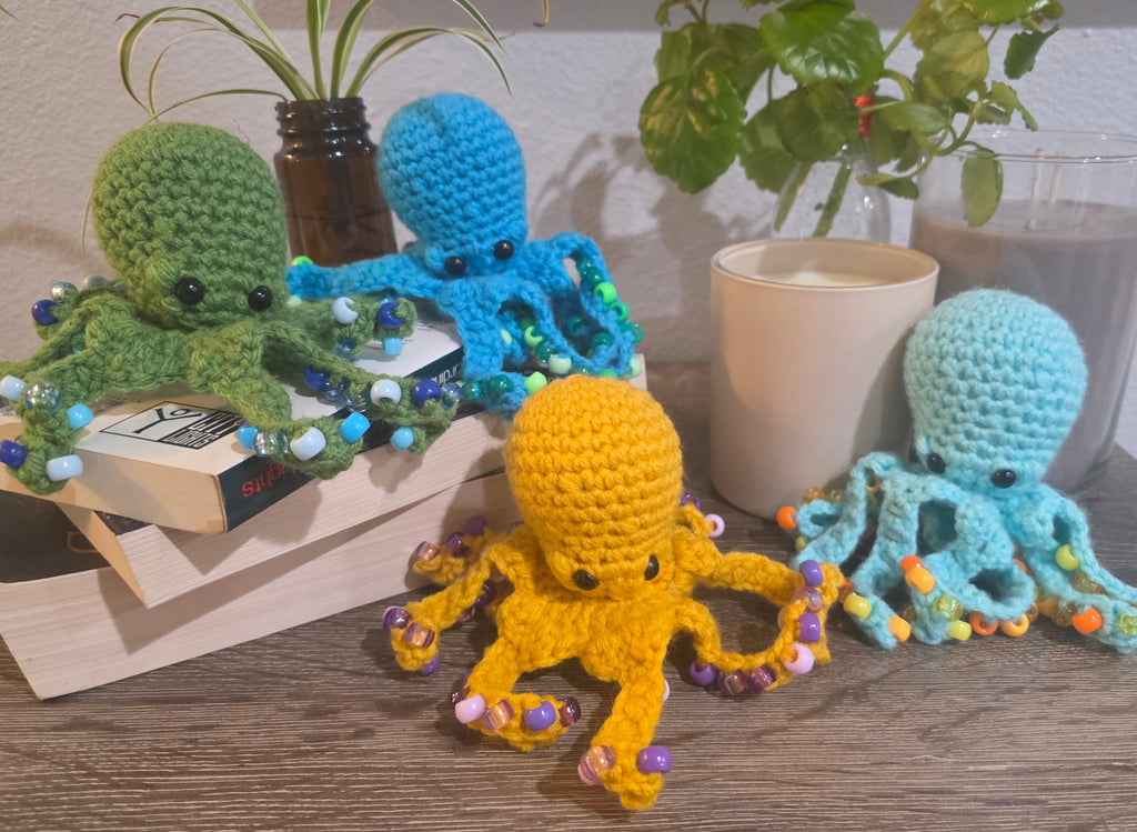 Four crocheted octopus toys in different colors on a surface with books and plants in the background.