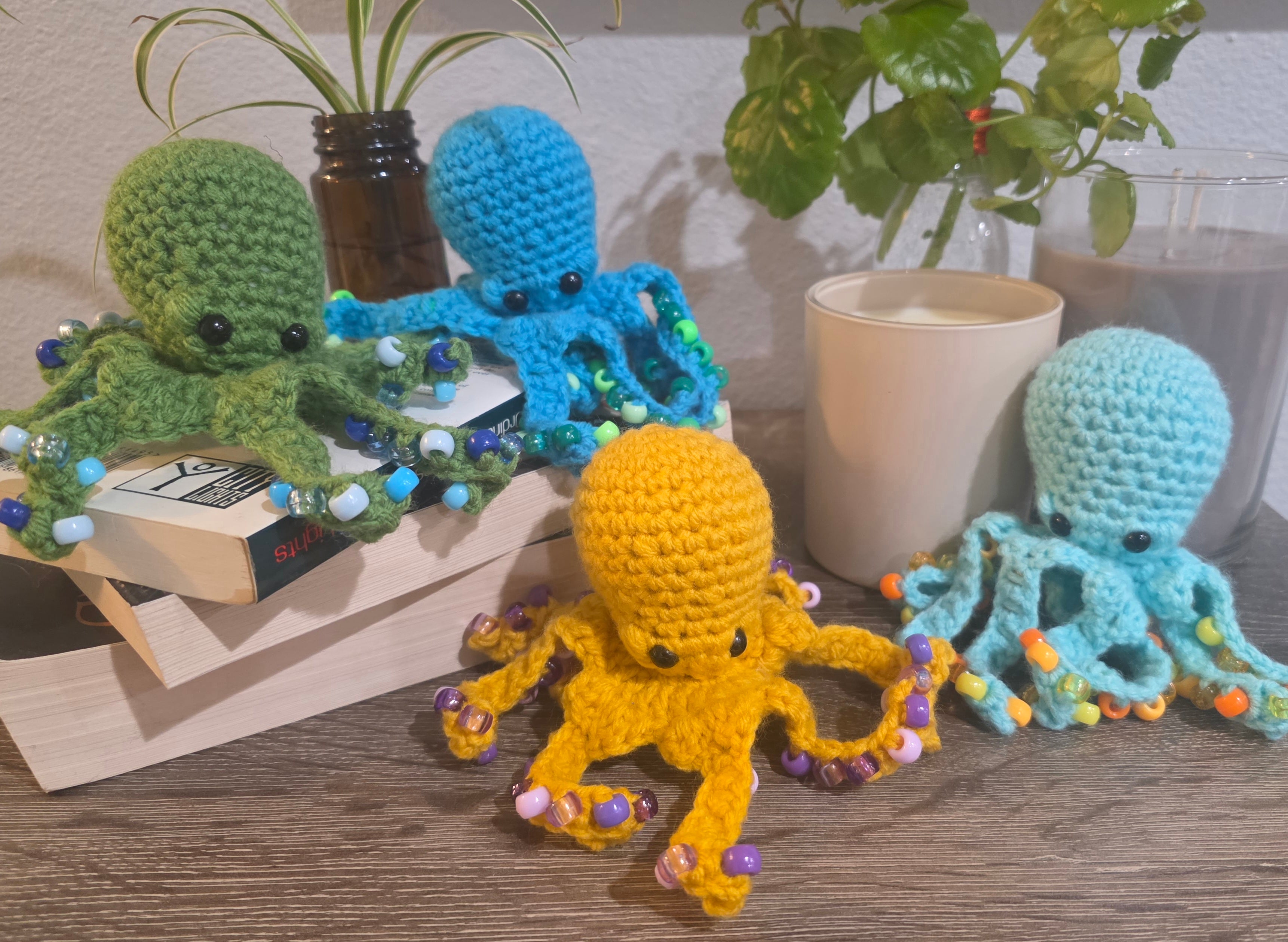 Four crocheted octopus toys in different colors on a surface with books and plants in the background.